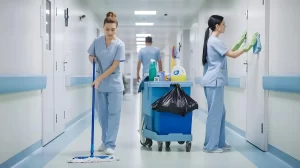 Profissional de limpeza hospitalar fazendo a higienização do chão com um esfregão, enquanto outra pessoa limpa as portas.