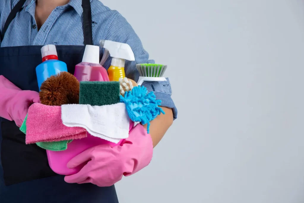 Profissional de limpeza e conservação segurando um balde com produtos e utensílios de limpeza, como esponjas e escovas.