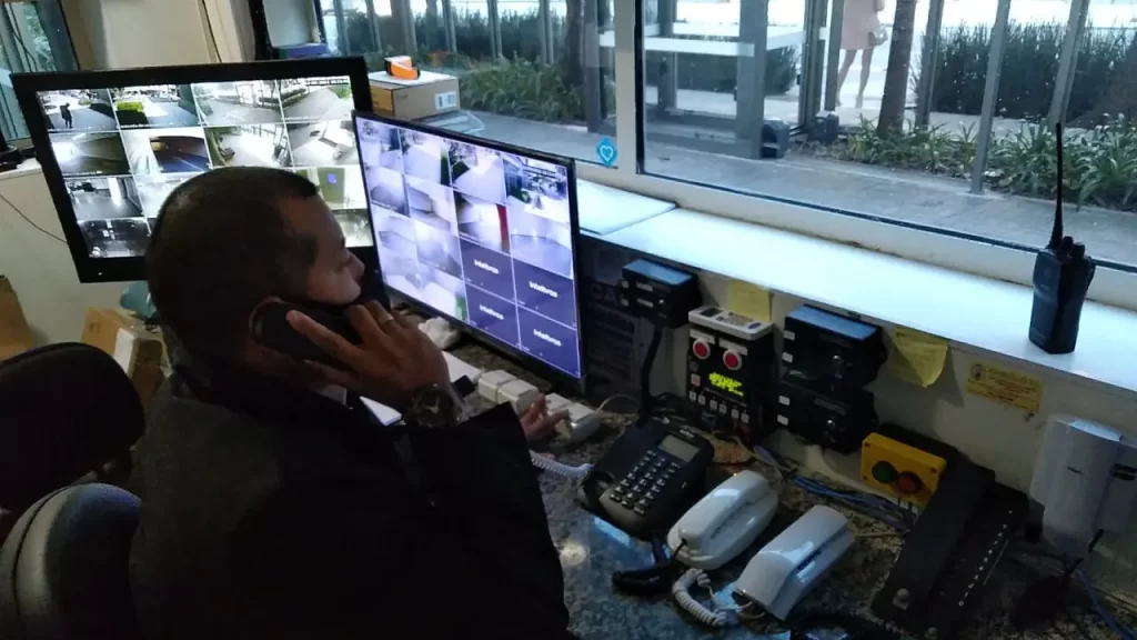 Trabalhador de portaria terceirizada falando ao telefone enquanto monitora câmeras de segurança de prédio.