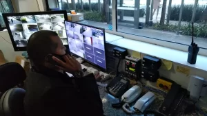 Trabalhador de portaria terceirizada falando ao telefone enquanto monitora câmeras de segurança de prédio.